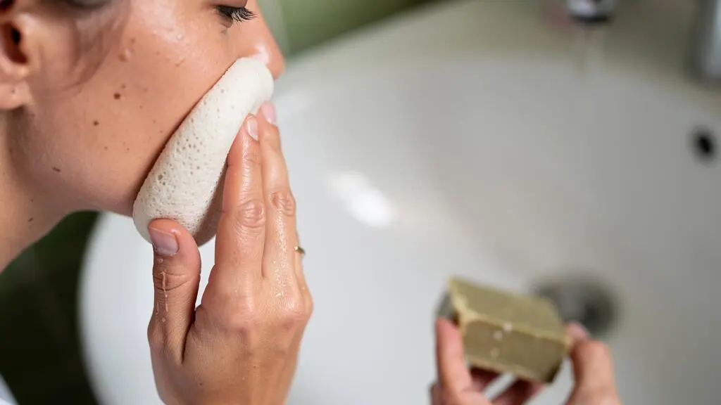 Des mains utilisent une éponge konjac humide et un pain démaquillant solide pour nettoyer un visage au-dessus d'un lavabo.